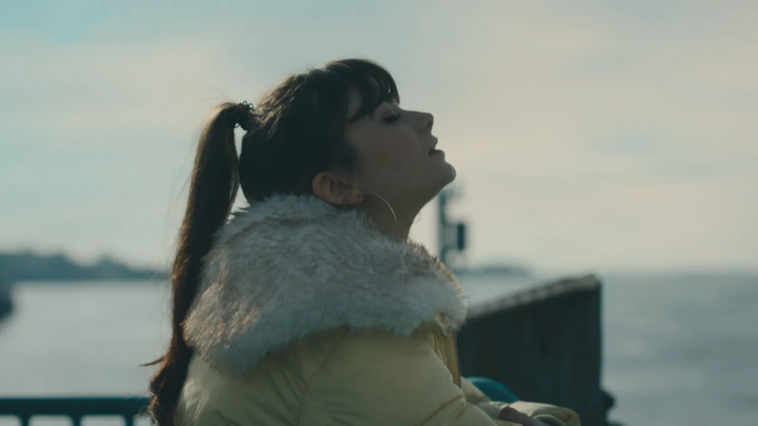 Tarian Cyf / S4C Effi wearing a yellow coat looks towards the sea with her eyes closed