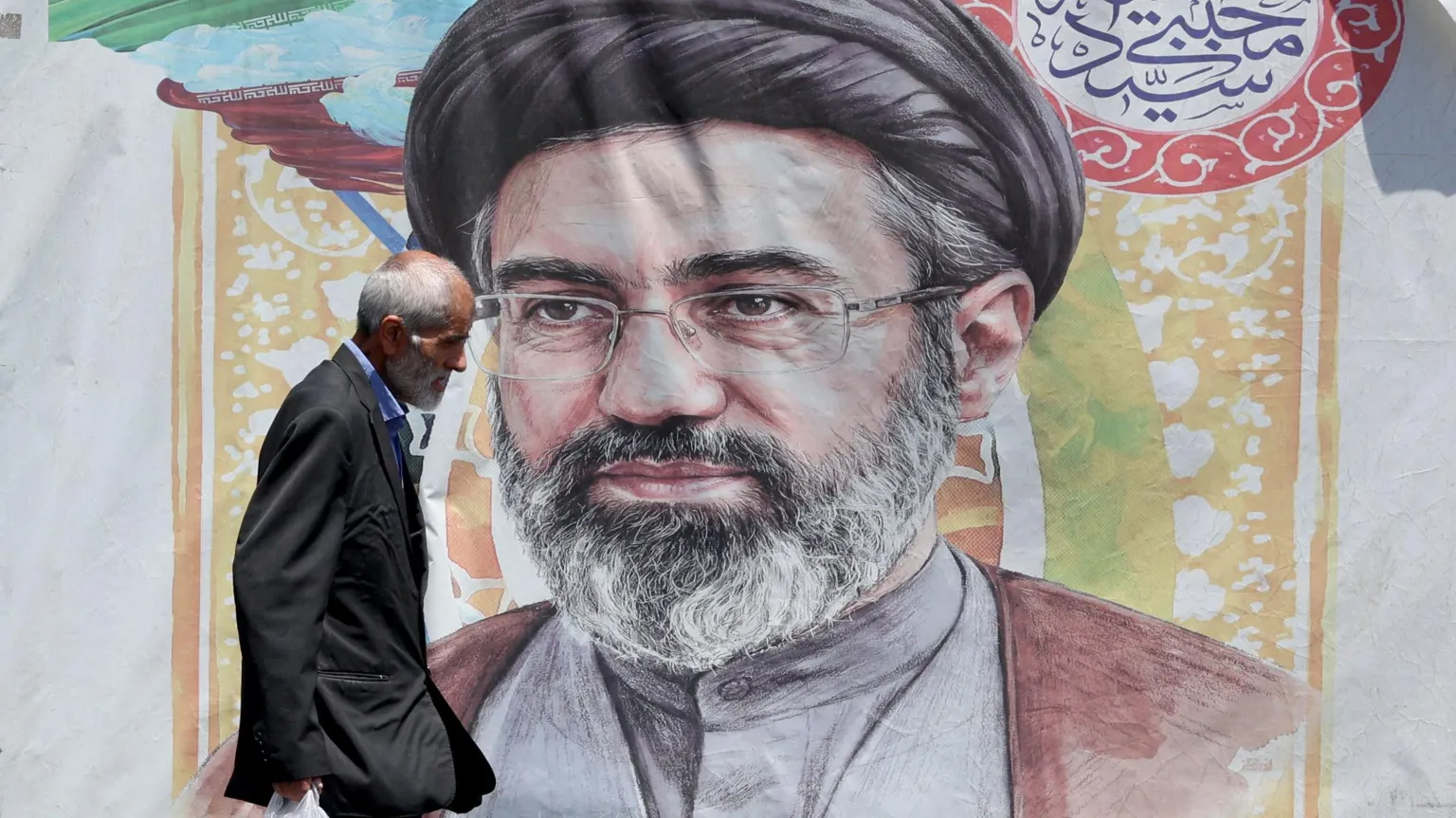 EPA A man with grey hair wearing a dark suit and blue shirt carrying a plastic bag walks past a poster of Supreme Leader Mojtaba Khamenei who has a grey beard and is wearing a grey buttoned shirt, maroon shawl glasses and a black turban.