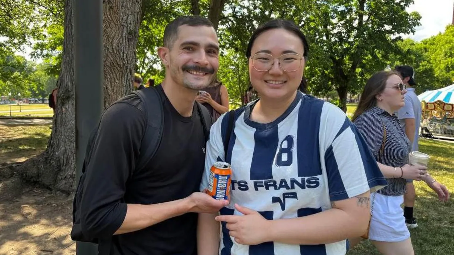 a man with short dark hair and a dark moustache standing next to a girl with her hair tied back and glasses. they are holding a blue and orange can of juice.