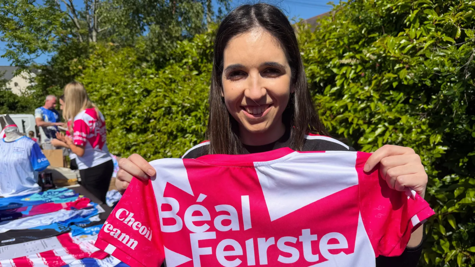 A woman, with long dark brown hair, holding a white and pink jersey reading