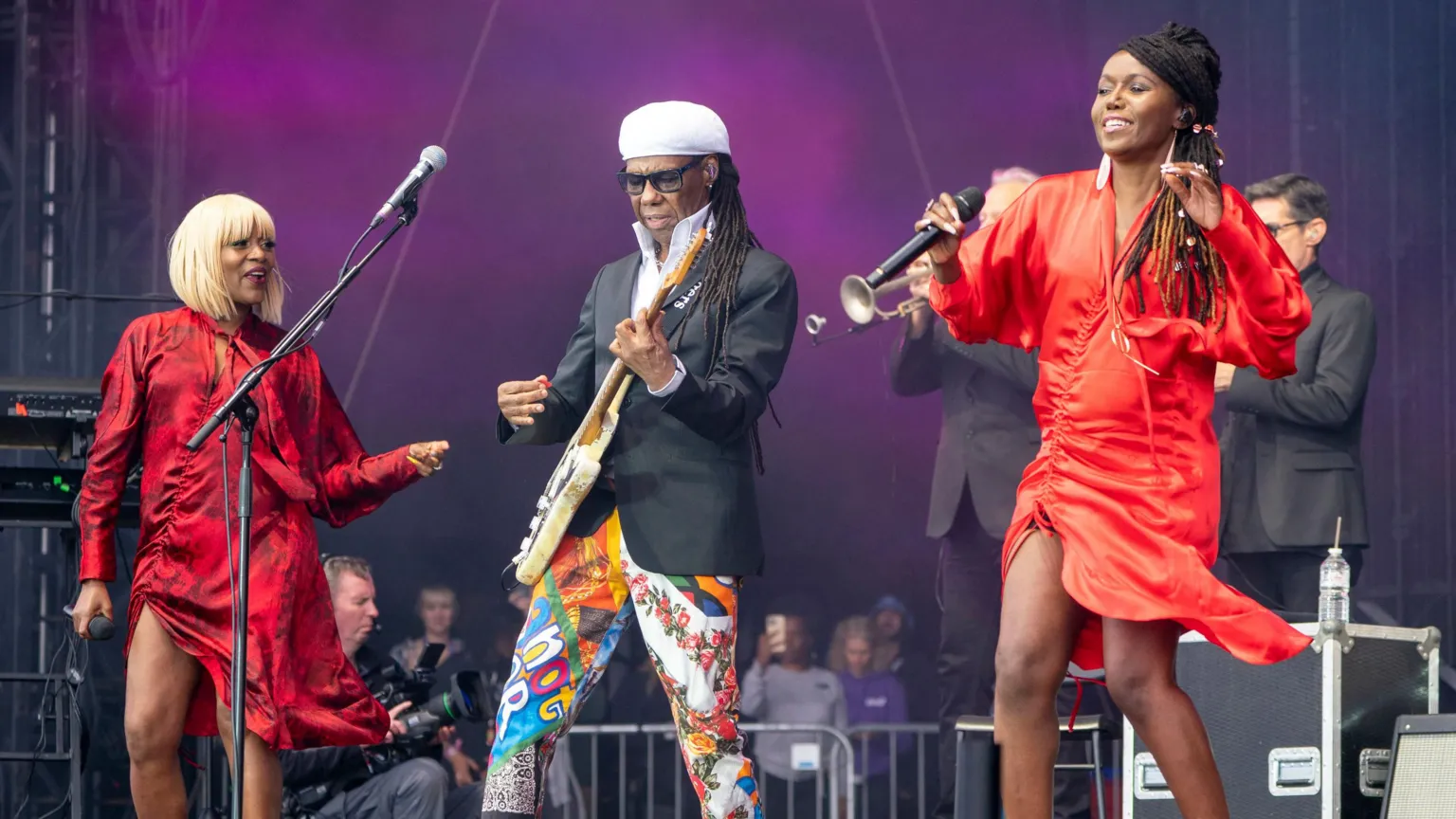  Nile Rodgers performs onstage at a music festival. He is playing guitar while multi-coloured trousers, a dark jacket and white hat, while two backing singers dance next to him, wearing red dresses.