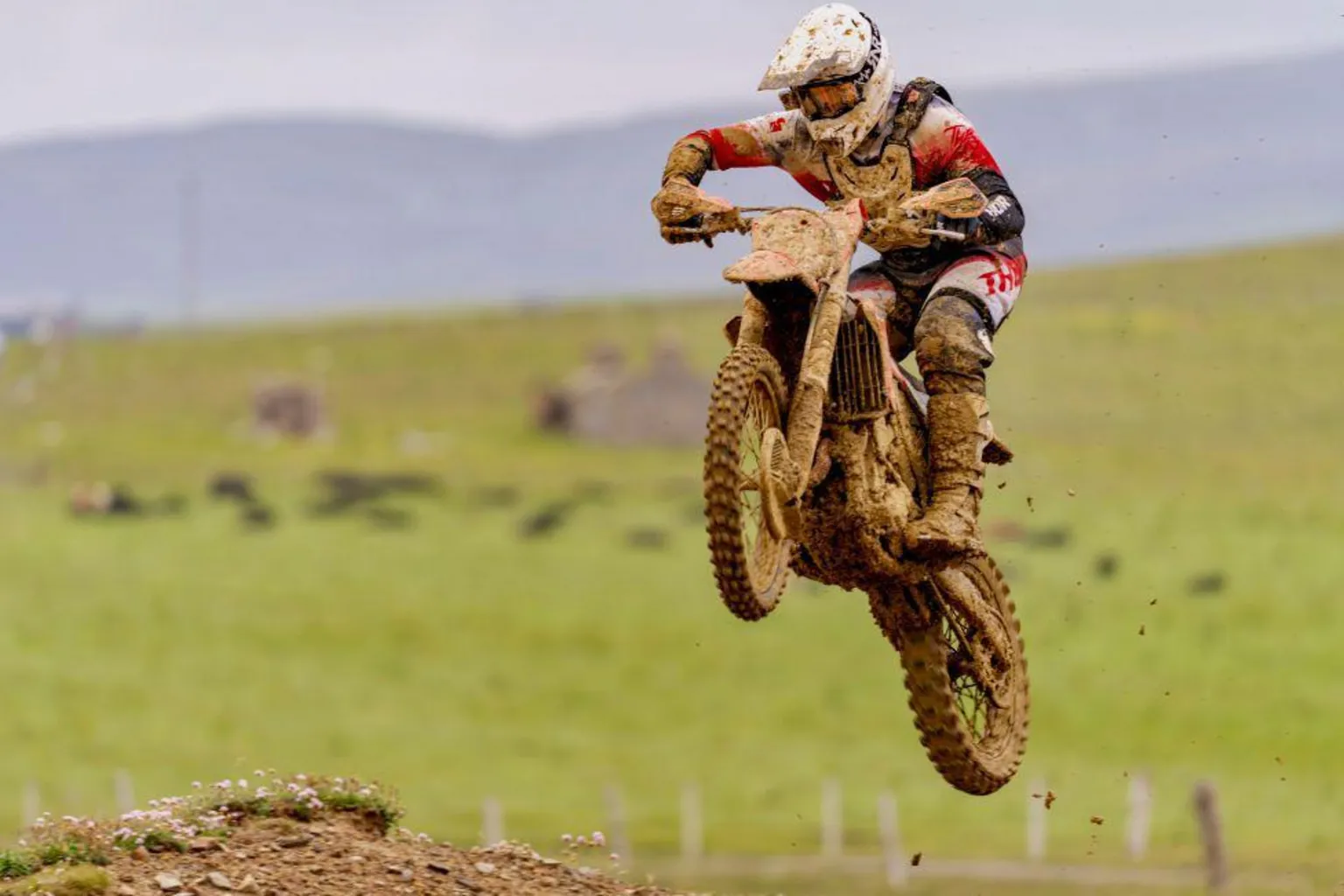 Bruce Carrington A motocross rider in a mid-air jump on bike, rider and bike are caked in mud, with a green field in the background.