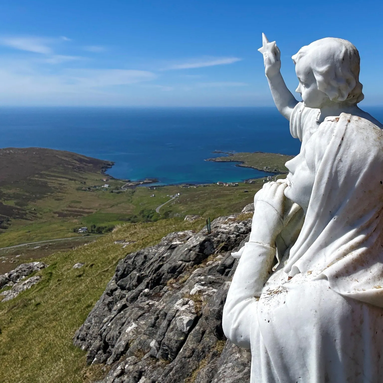 Laura Docherty A white statue of a woman and child high on a hillside, overlooking the sea, under a blue sky.
