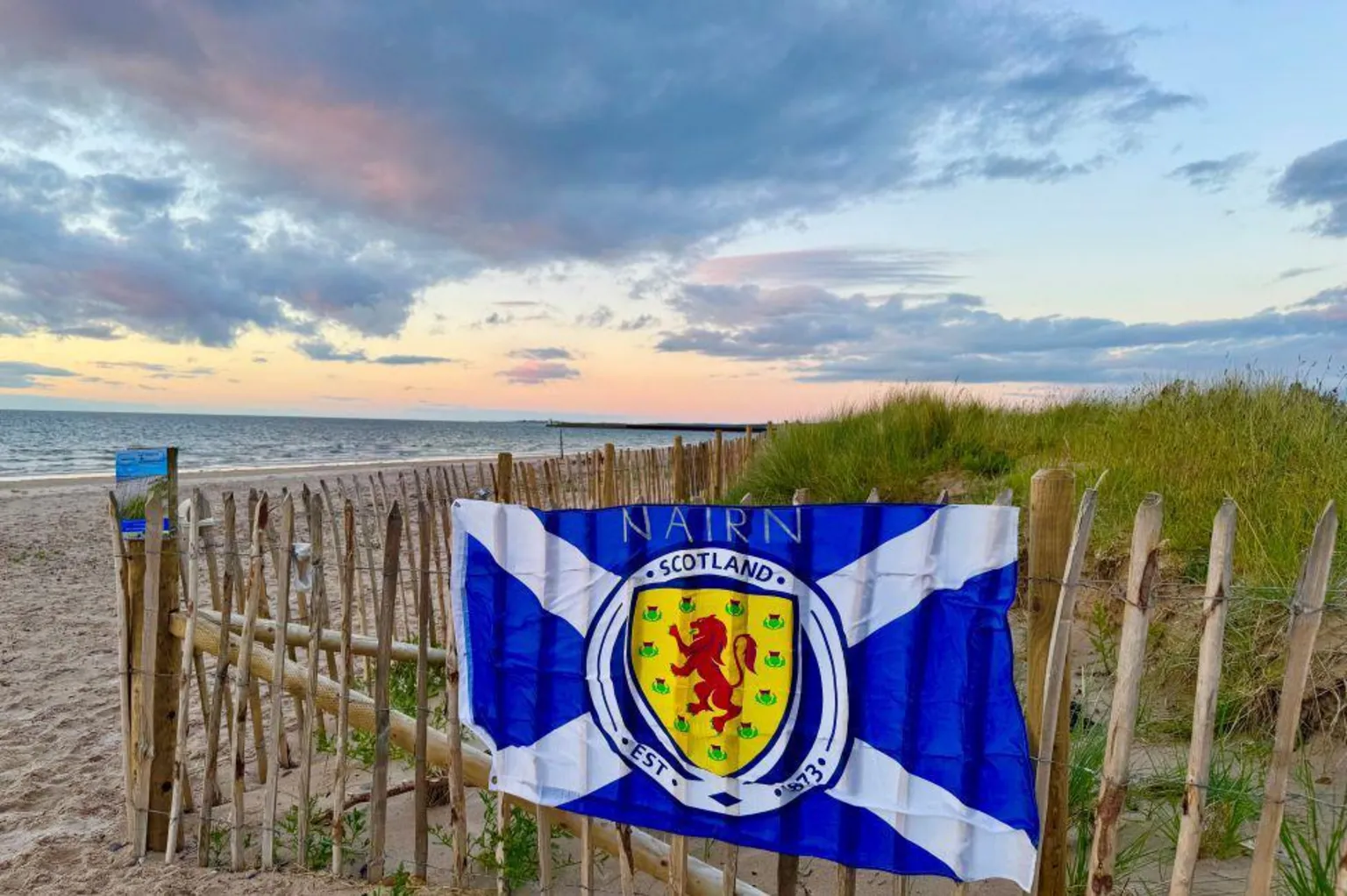 Elena Reid Saltire flag pictured at sunset on Nairn beach, hung up on a fence, with beach and pastel sunset sky in background.