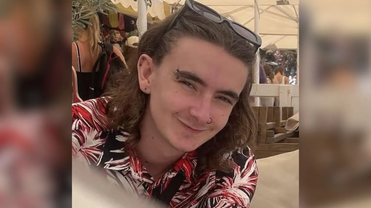 Family Handout Jamie Collins wearing a black, red and white patterned shirt and sunglasses on his head. He has shoulder length brown hair and piercings in his ears, eyebrow and nose. He appears to be sitting underneath a white marquee on a sunny day.