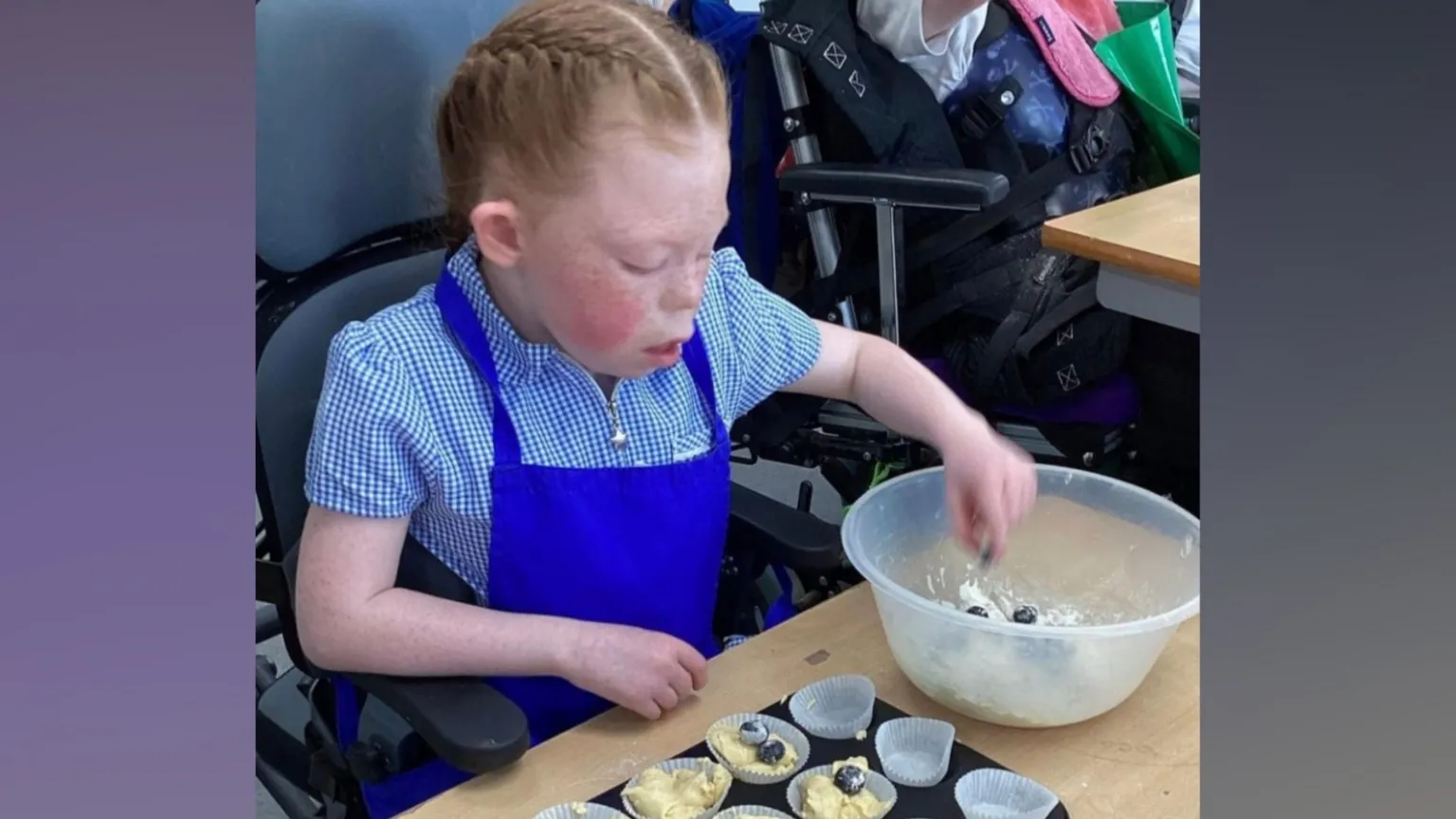 Ridgeway School Amelia sits at a desk in school wearing her checked blue dress and a royal blue apron. She is making cakes and has her hand in a clear plastic bowl, picking out blueberries. Her auburn hair is tied back in a French plait and her cheek is flushed.