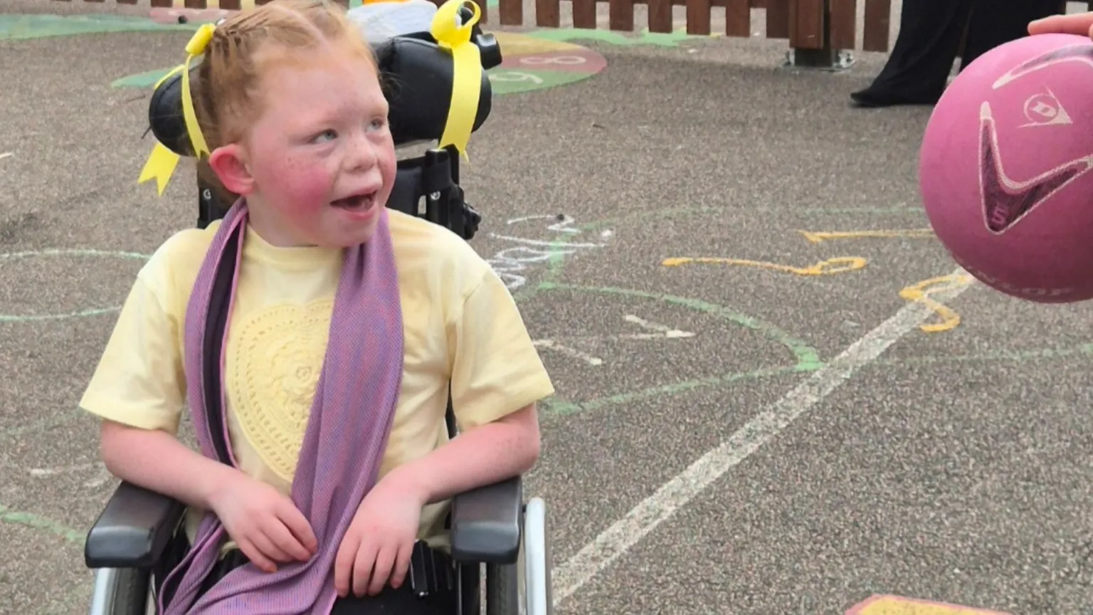 Ridgeway School Amelia sits in her wheelchair outdoors wearing a cooling towel around her neck. She is looking up at someone off camera who is holding a football. She has yellow ribbons in her hair and a yellow T-shirt.
