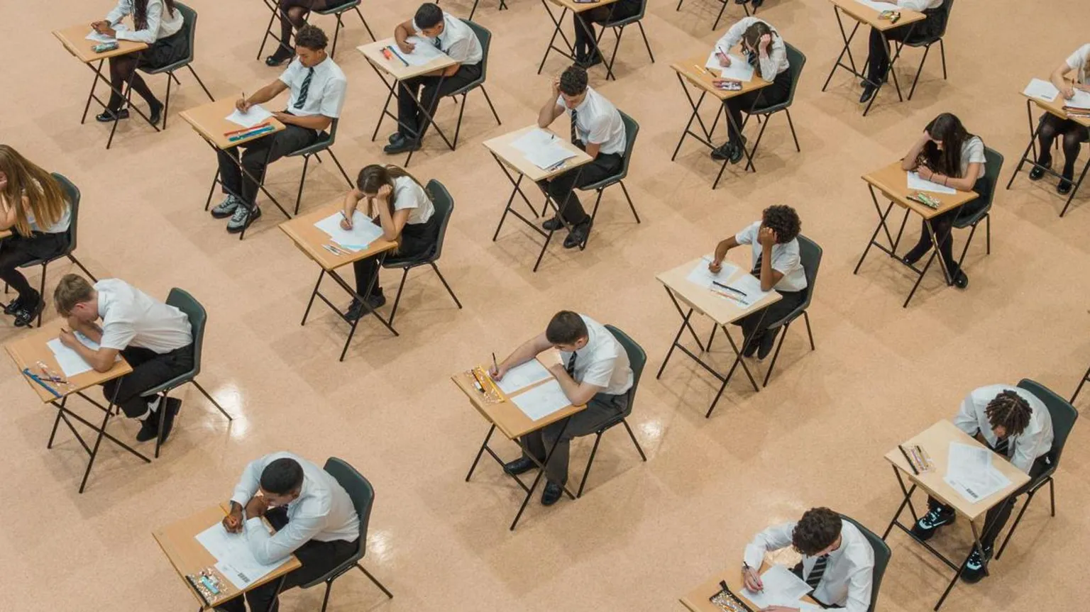 Students sitting their exams, taken from above in an aerial view. They are wearing shirts and ties and sitting at individual desks, writing their exam papers.