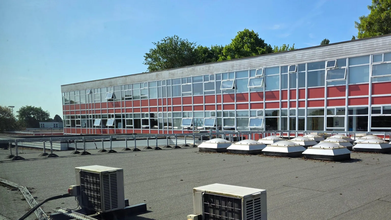 Sele School A photo of the exterior of the Sele School shows the top floors, which are almost entirely made up of windows, some of which are open. Part of the building below that is a flat roof with some heating/air units on. The sky is blue.