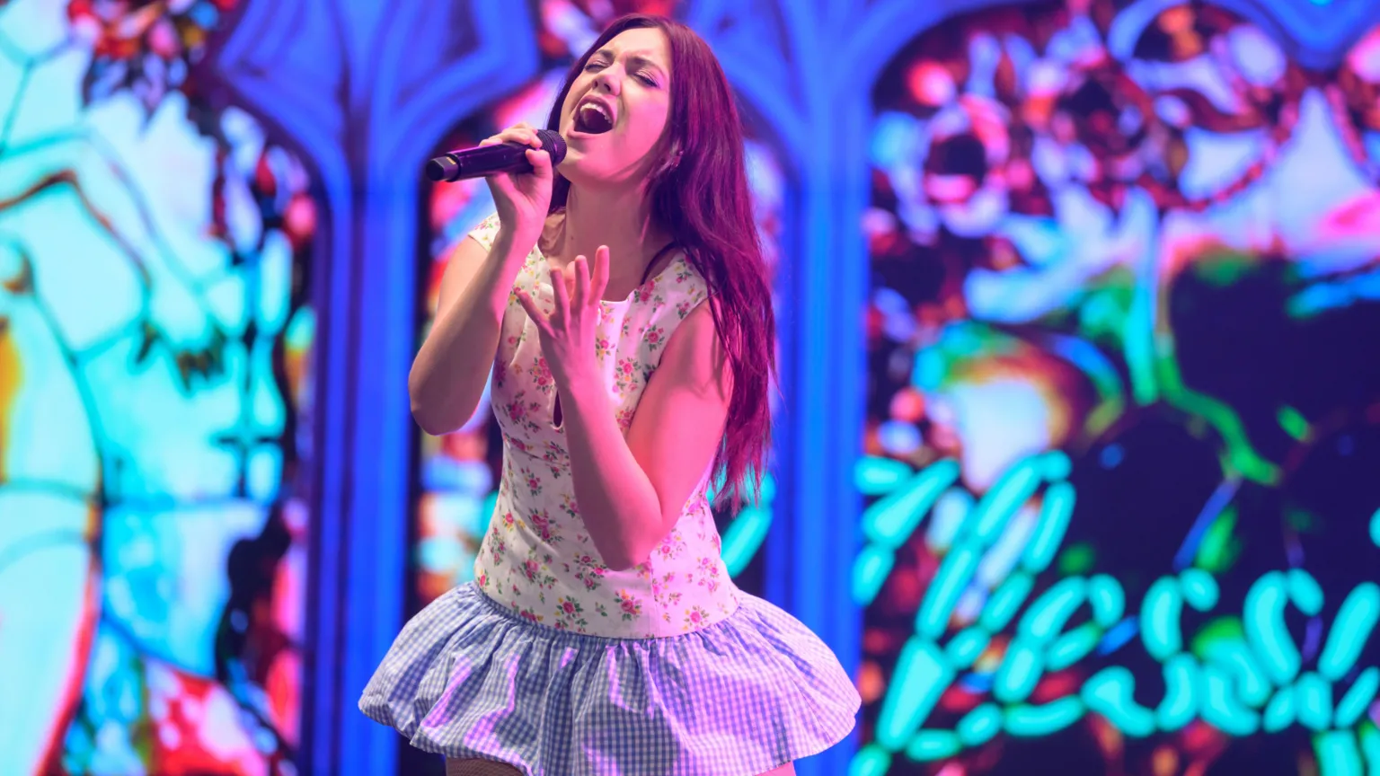 Alessi Rose is singing in to a microphone. She is on a stage which has a stainglass affect background. She wears a mini-dress which has a floral print top and a blue and white gingham-style skirt.