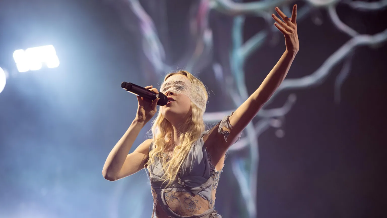 Erin Le Count performs on stage and is holding a microphone. She is wearing a blue dress with lace cut-outs and has a lace eye-covering.
