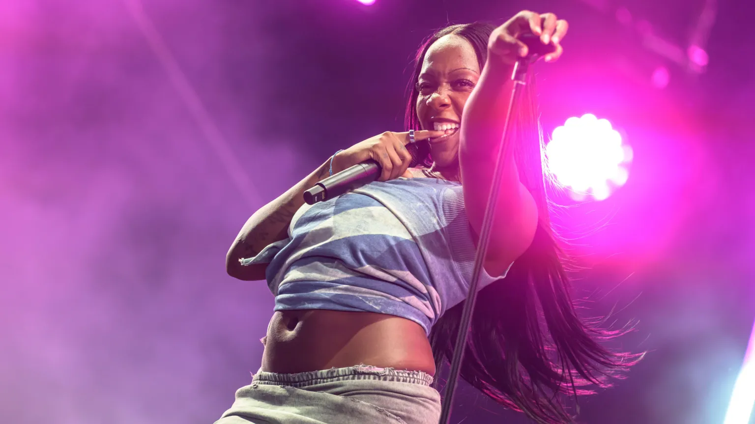 Rachel Chinouriri performs on stage. She is holding a microphone stand in one hand and a microphone in the other while biting her finger. She is wearing a blue cropped top and grey skirt. She has long brunette hair.
