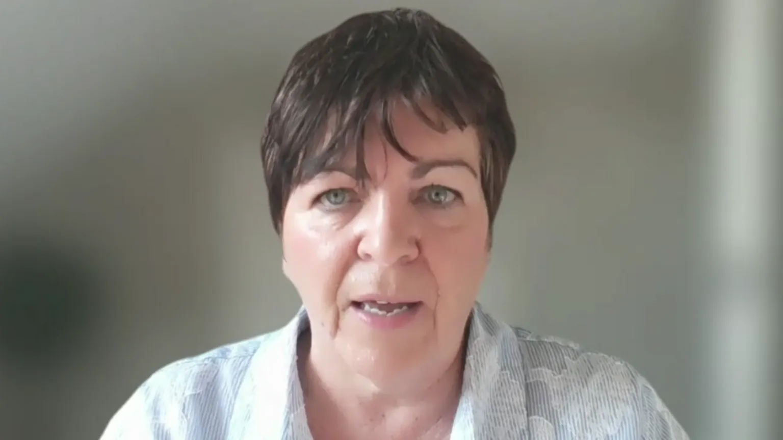 I woman with dark hair is speaking directly to the camera. She has short dark hair and is wearing a pale blue and white shirt with applique flowers. 