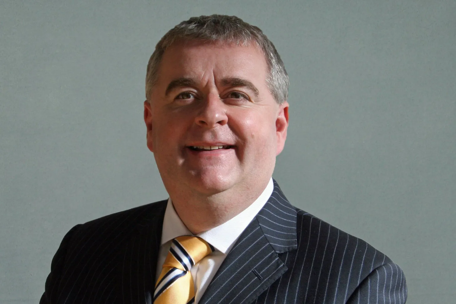SBC A man with dark hair, wearing a dark jacket, white shirt and yellow tie looks at the camera