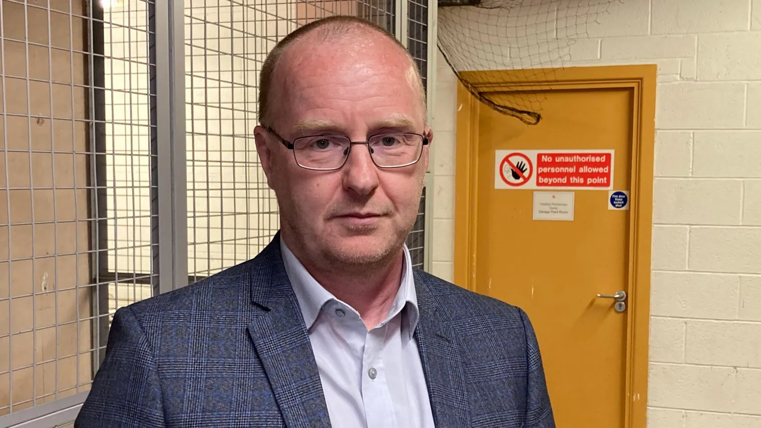 A man with shaved hair and rectangular rimmed glasses looks at the camera. He wears a grey cheque suit jacket and has his hands in his trouser pockets.