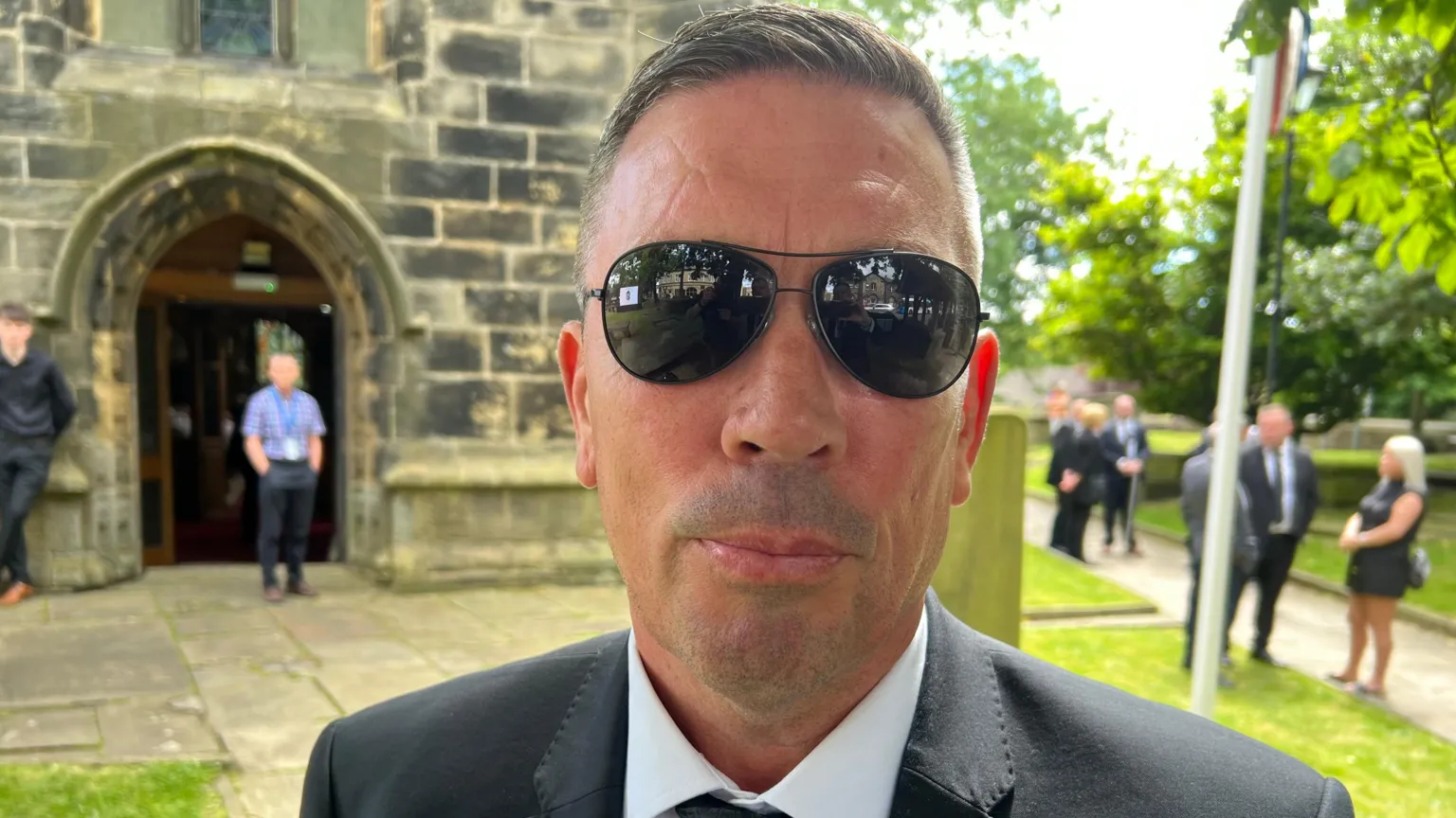 Paul Ogden/BBC A man with short grey hair wearing black sunglasses looks at the lens. Behind him is a church and other people dressed in black. He has a black suit jacket on.