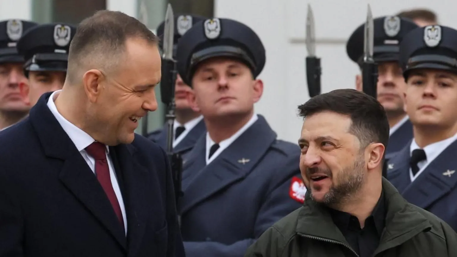  Polish President Karol Nawrocki and Ukrainian President Volodymyr Zelensky smile at each other while soldiers stand to attention behind them.