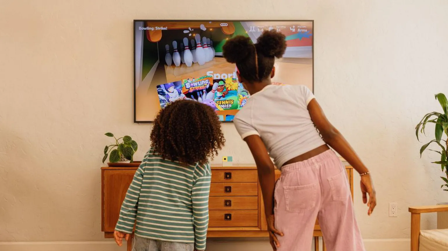 Nex Playground Two children are staring with their backs to the camera in a living room space facing the TV screen which is showing a bowling game. Beneath the screen the Nex Playground console cube in white, green and yellow sits on a table.