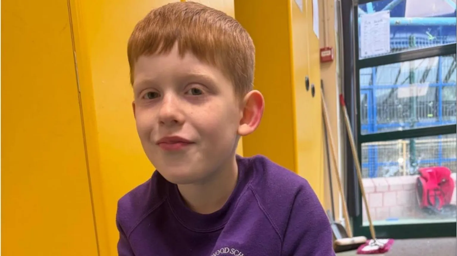 Amy Gibney A young boy with ginger hair sits in front of a yellow wall, wearing a purple jumper. Over his left shoulder is a clear window with blue railing behind it.