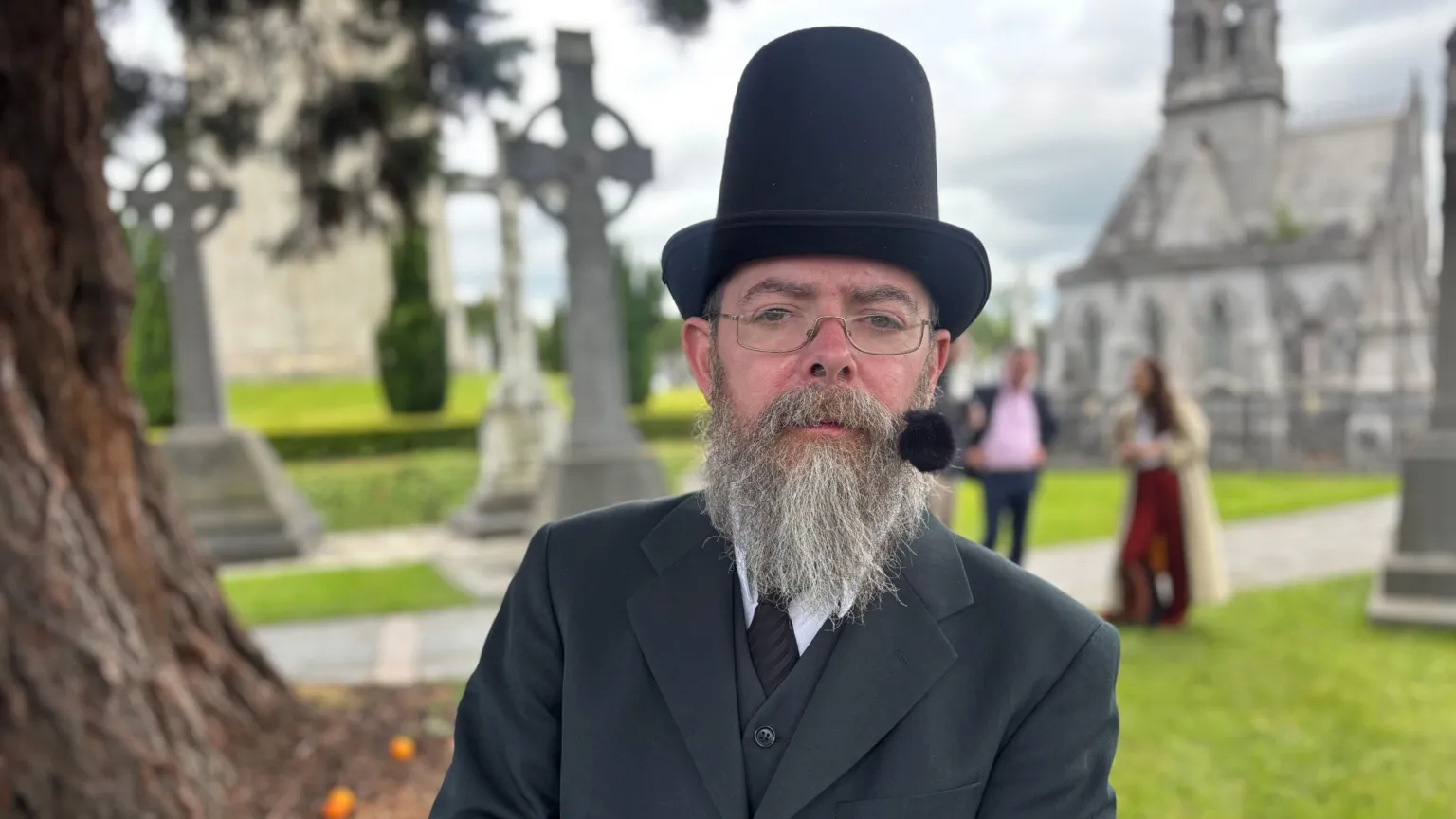 A man wearing a black three piece suit and tie and a black tophat, with a grey long beard, is photographed at Glasnevin Cemetery 