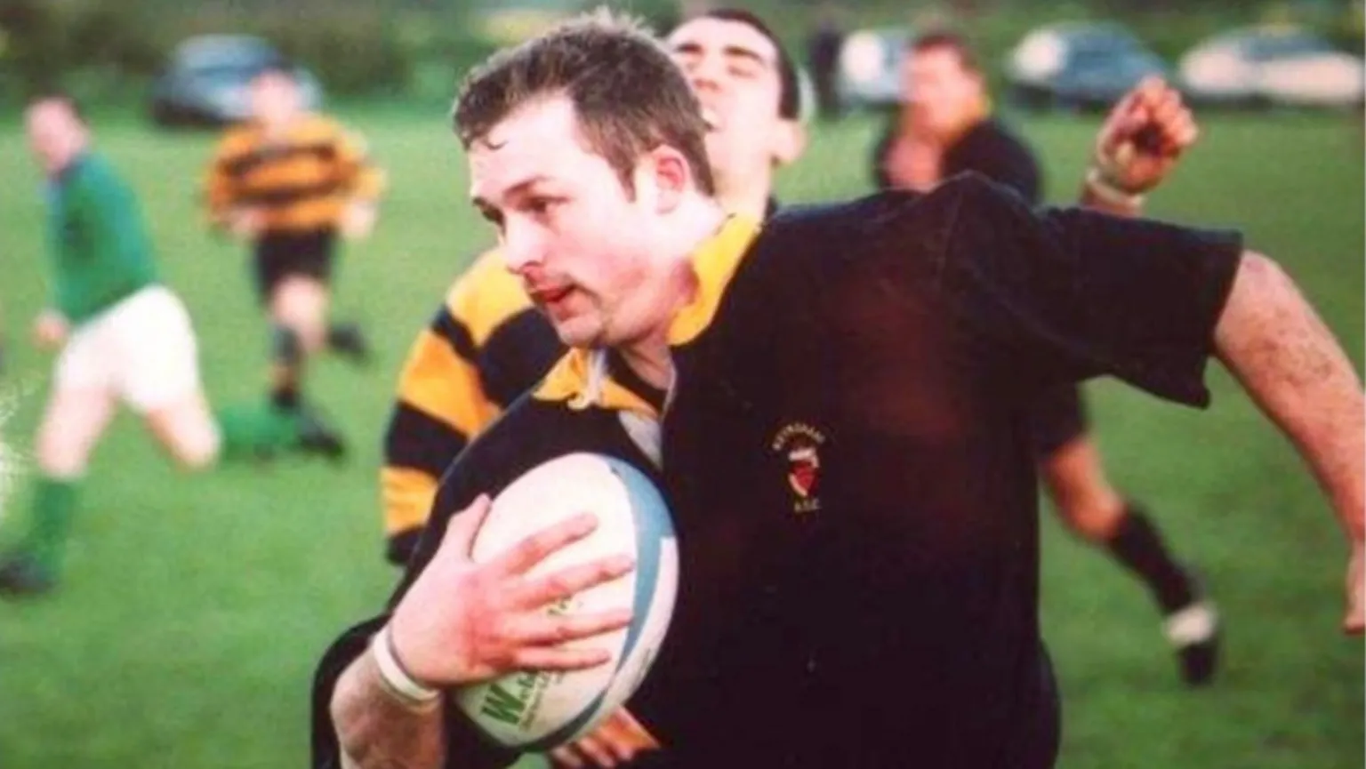 Family photo/Keynsham RFC A man with brown hair wearing a black and yellow rugby strip and running on a field holding a rugby ball. Other players are visible in the background.