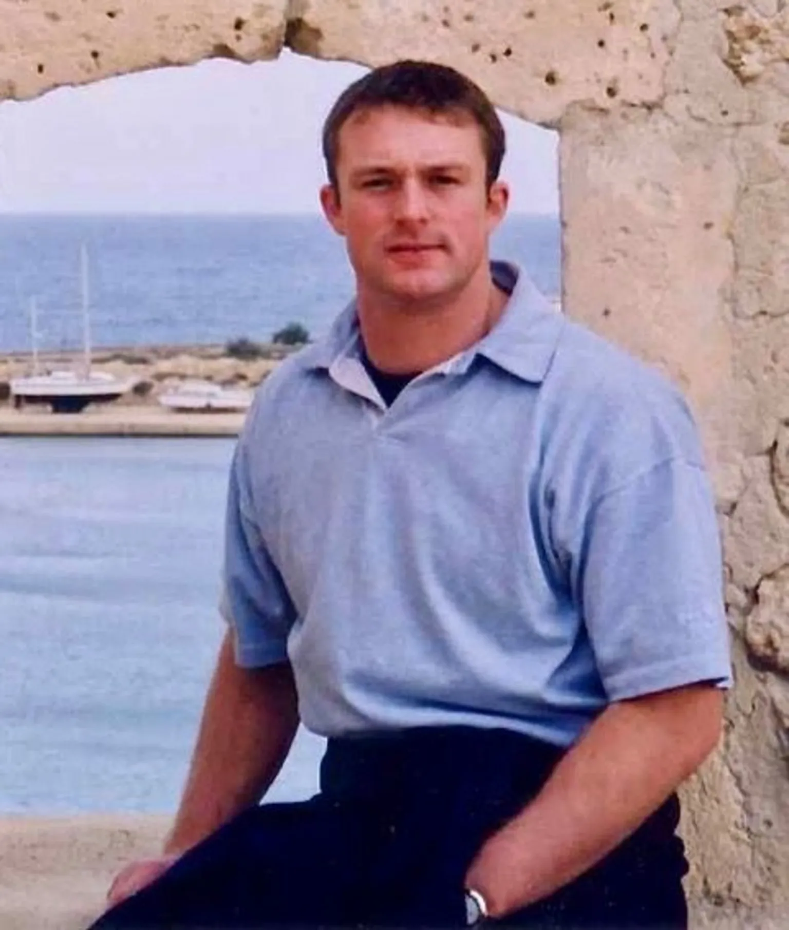 Family photo A man with brown hair, wearing a blue polo shirt and dark trousers. He is sat on a stone ledge with a stone wall and sea visible in the background. He has one hand in his pocket.