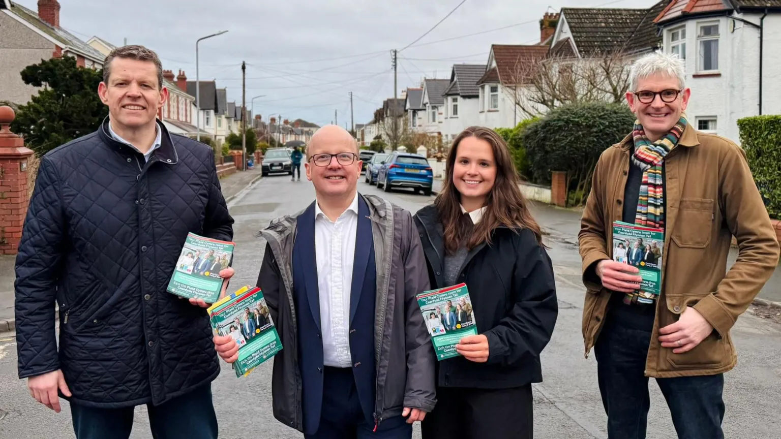 Plaid Cymru Plaid Cymru elected members campaigning on a street in Wales. Plaid Cymru party leader Rhun ap Iorwerth is stood on the left of the image. Stood next to him is Dafydd Trystan Davies. Next to Dafydd Trystan Davies is Zaynub Akbar. On the far right of the image is Nick Carter. Each elected memeber is holding campaign pamphlet's. Cars and houses can be seen behind them.