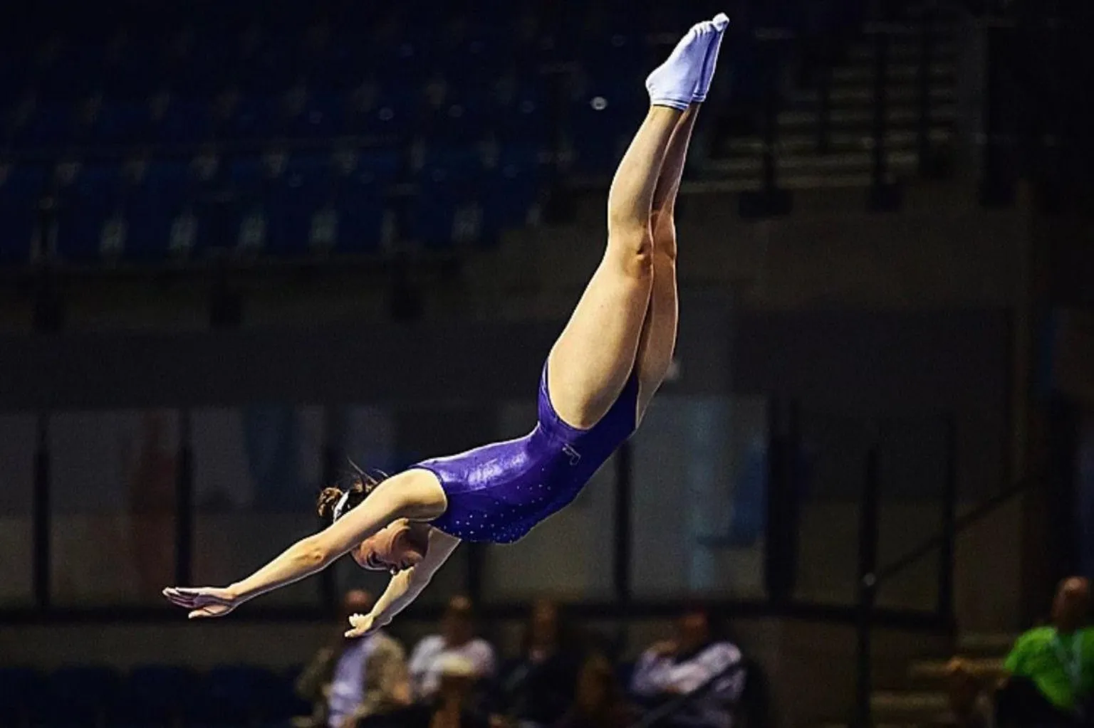 British Gymnastics Akbar competing in gymnastics in mid-air arms outstretched in purple leotard