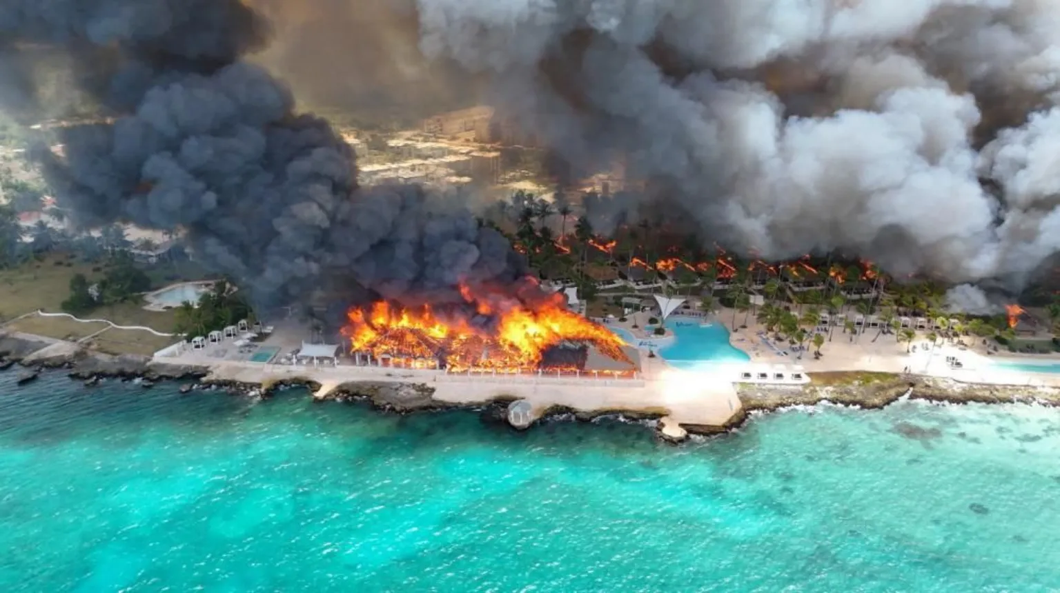  Aerial shot showing a large building on fire, and thick black smoke. The resort is on the beach and the water is vibrant blue.