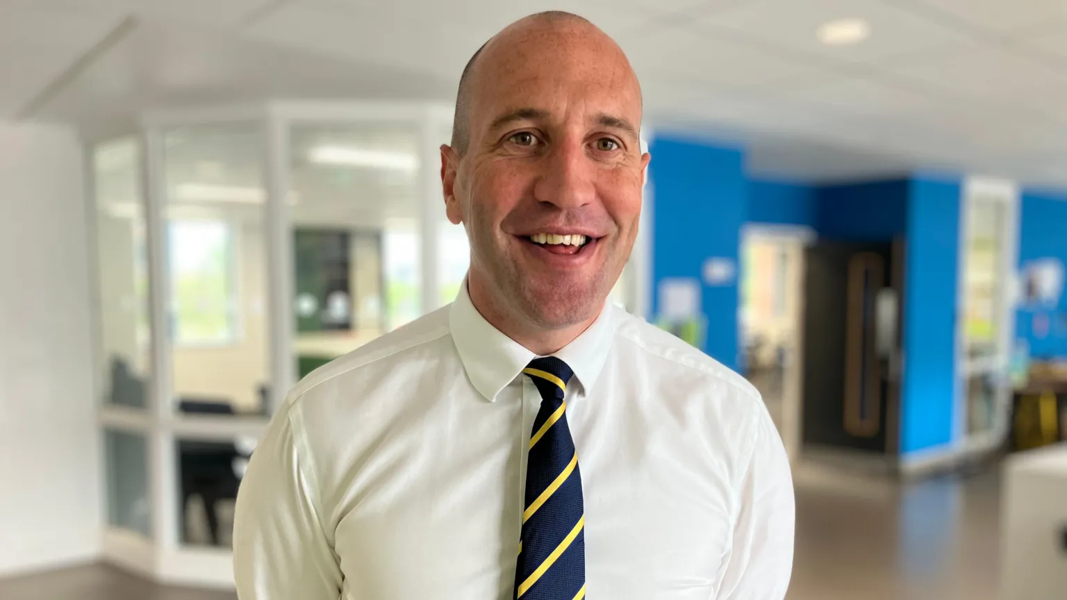 Innes Robinson is wearing a white shirt and navy and yellow striped tie, he is standing in his school canteen.