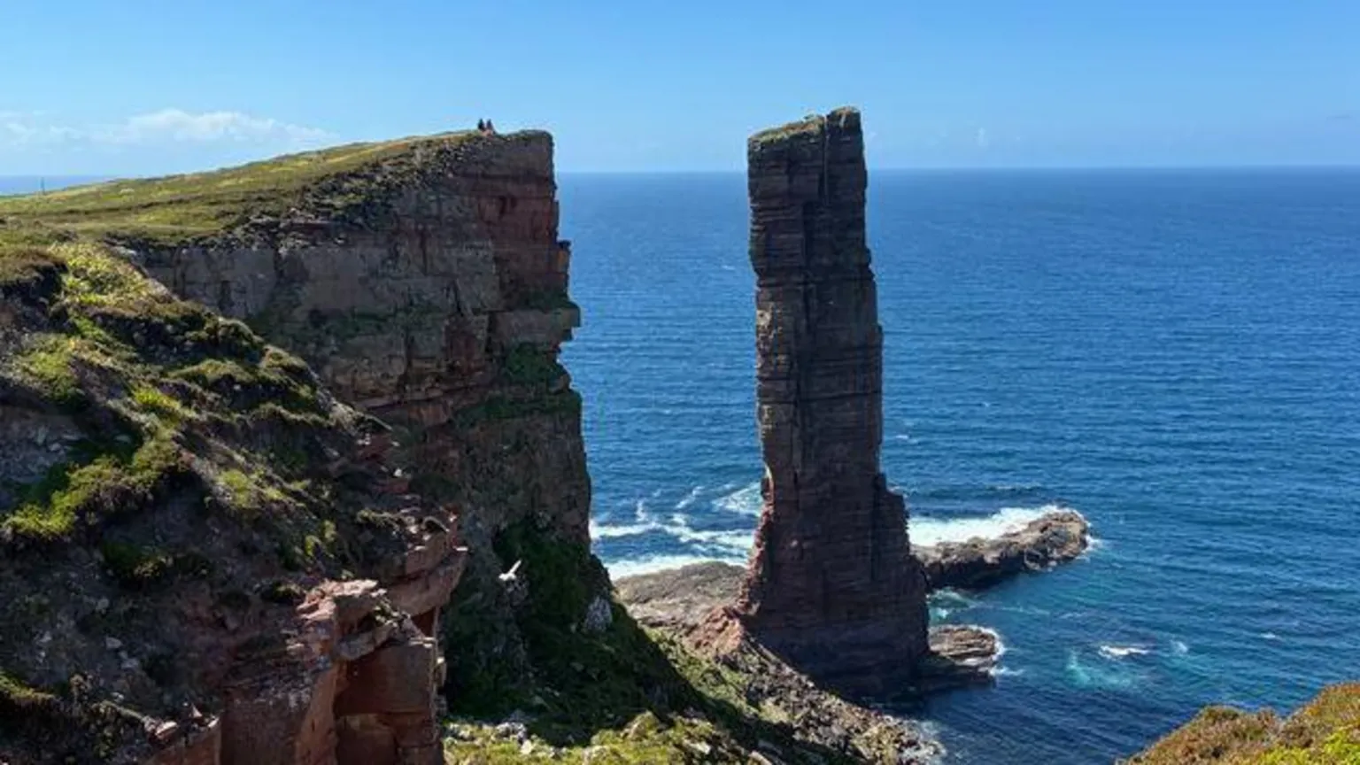 Two figures sit on a cliffside overlooking the Old Man of Hoy, an iconic sea stack in Orkney.