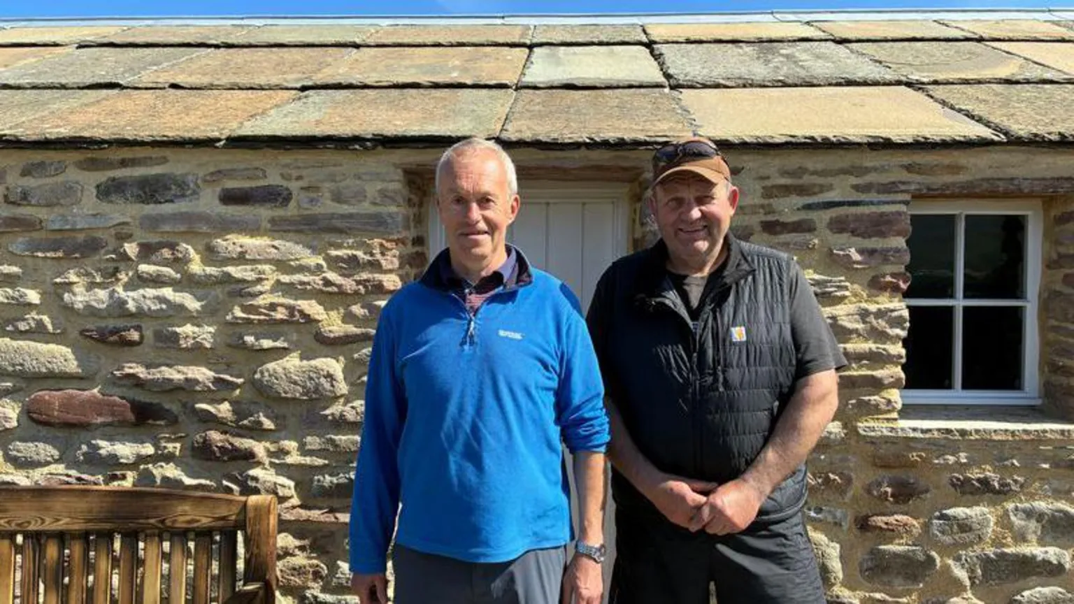 A man in a blue fleece stands next to a man dressed in a black gilet and shorts in front of the newly restored bothy. It is a sunny day, and they are smiling.