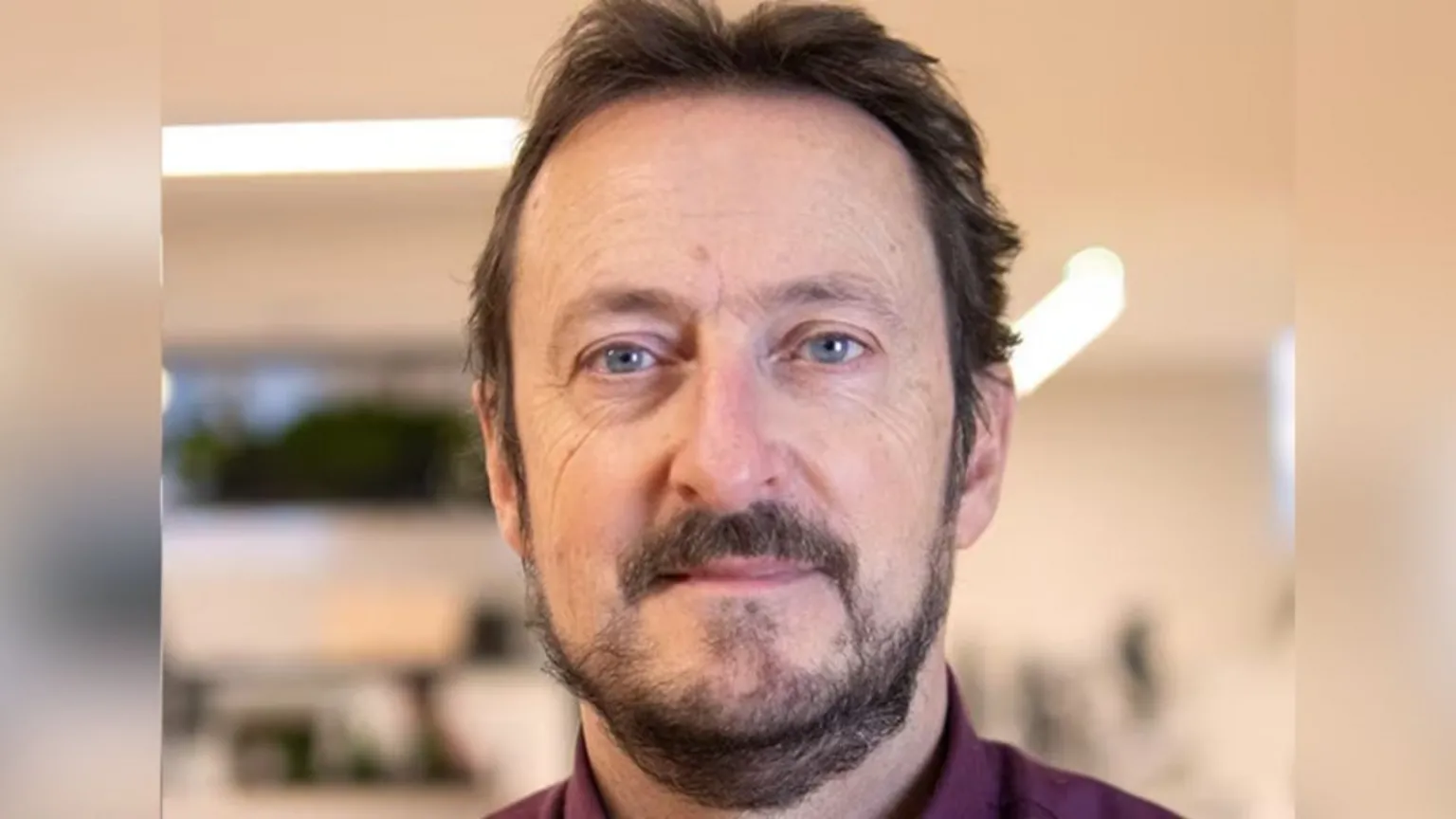 Neil Johnston A close-up portrait of Neil Johnston with short dark hair and a trimmed beard, looking at the camera indoors, with a softly blurred background showing shelves and overhead lighting.