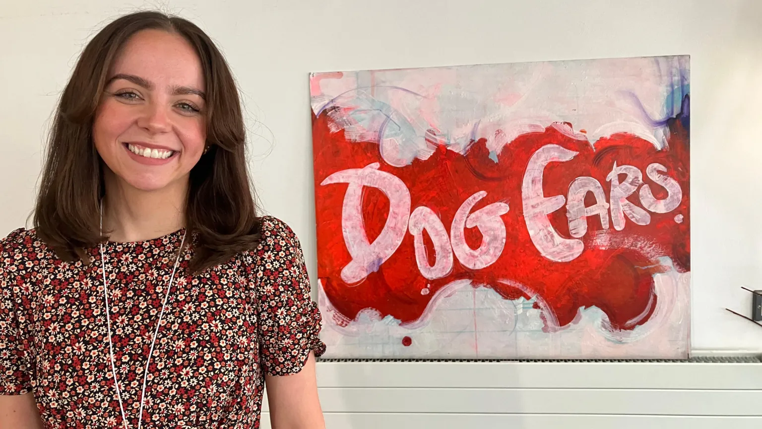 A woman with shoulder length brown hair, wearing a floral pattern top. She is smiling at the camera. There is a paining on the wall beside her with the words