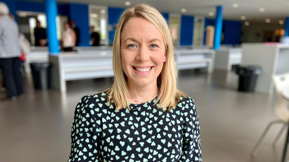 Mae Hayley Prosser yn eistedd yn ffreutur yr ysgol. Mae hi'n gwisgo ffrog dywyll gyda chalonnau gwyrdd.