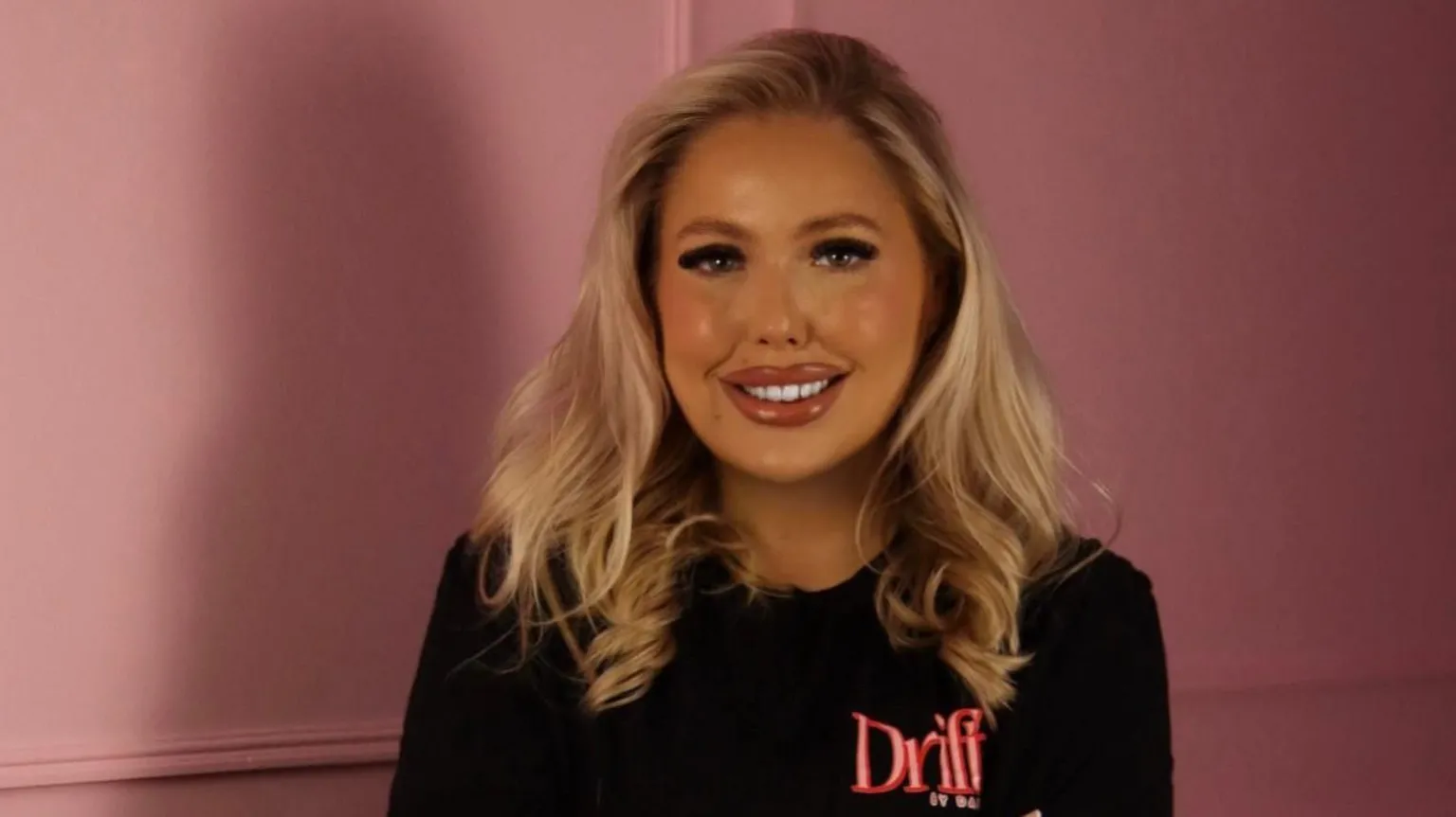 Close-up studio-style portrait of Dani wth shoulder-length blonde hair wearing a black top with the word “Drift” in pink, seated in front of a pink wall with soft lighting.
