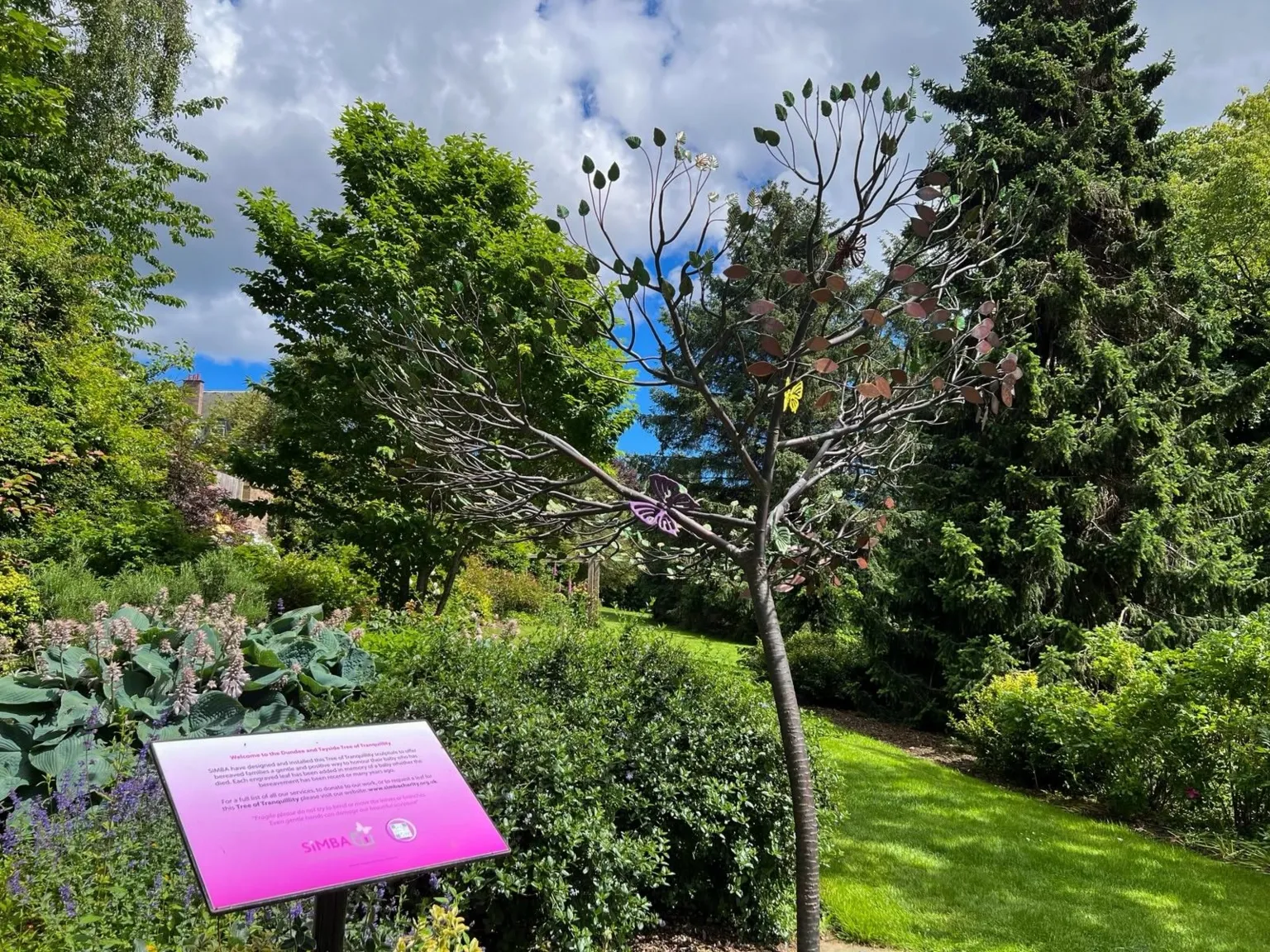 A metal tree sculpture with copper leaves sits in a large garden. A pink sign describing the tree is in the foreground