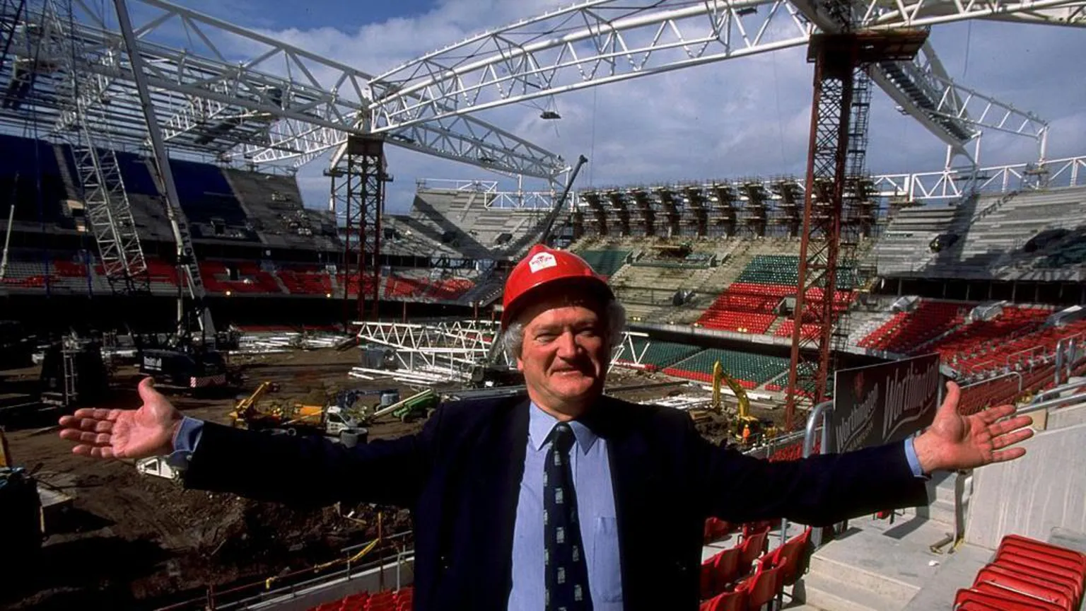 Glanmor Griffiths in a red hard hat, light blue shirt, dark jacket and WRU tie. He faces the camera, in the background are cranes and diggers during the early stages of the Millennium Stadium's construction