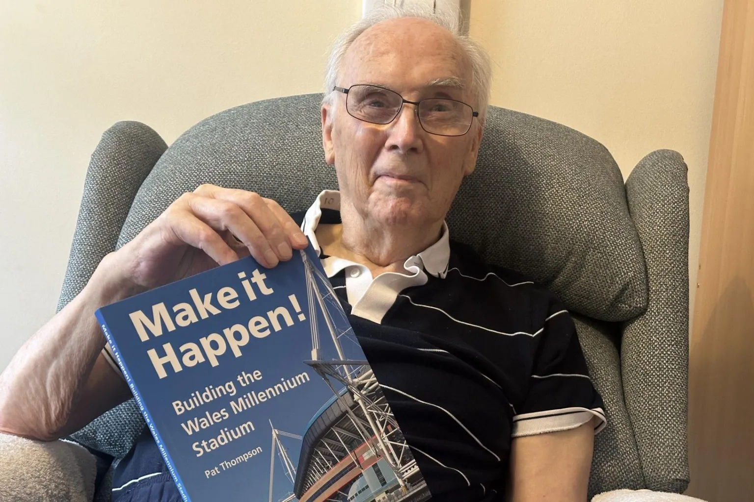 An elderly Pat sits in an armchair with a copy of his book. He has white hair and spectacles and is wearing a navy polo shirt with a white collar