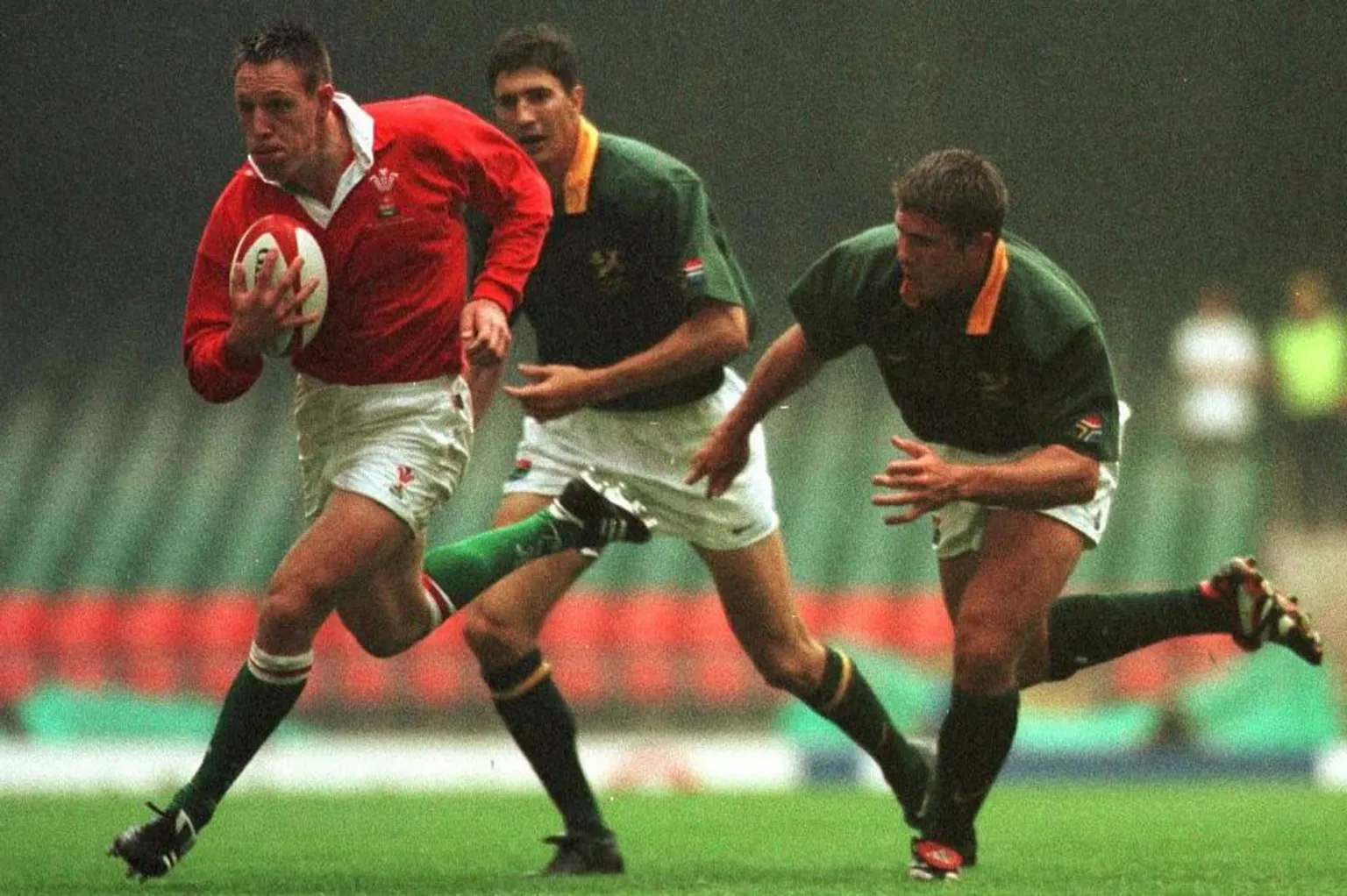 Mark Taylor charges towards the tryline, ball-in-hand, with a determined look on his face. Chasing him are two South African players, dressed in white shorts and green shirt with gold collar