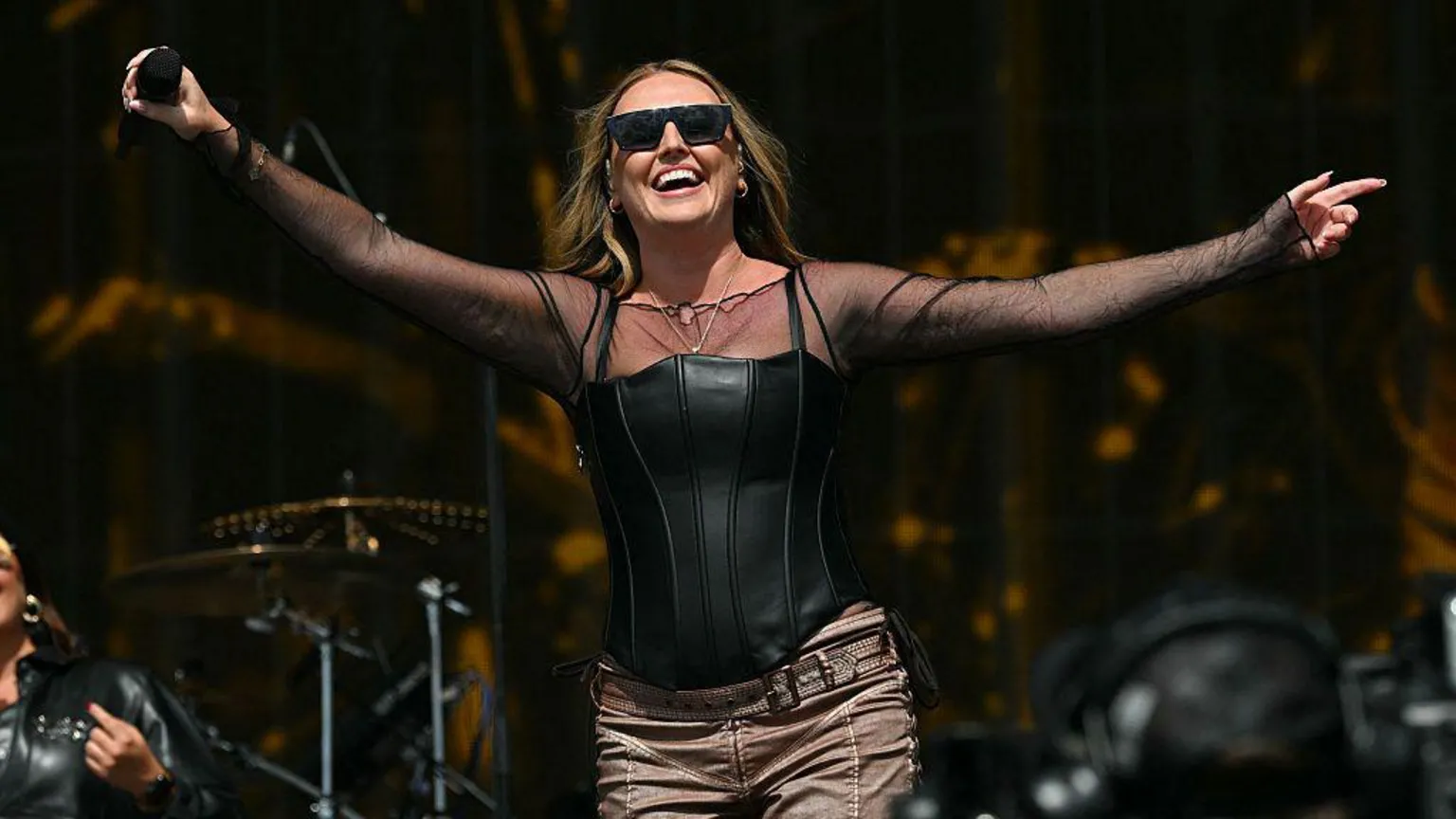  Perrie performs on stage at the TRNSMT festiva. She is holding a microphone and smiling, wearing a black top and with shoulder length brunette hair.