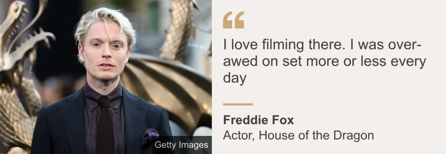  Freddie Fox in dark suit standing in front of a golden statue