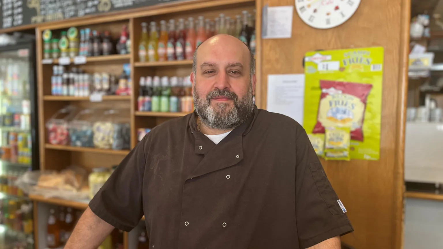 Adriano Pia with a shaved head and a full beard wearing a dark chef-style jacket, standing behind a counter in a shop with shelves of bottled drinks and snacks, a wall clock, and a “Savoury Treats” display visible in the background.