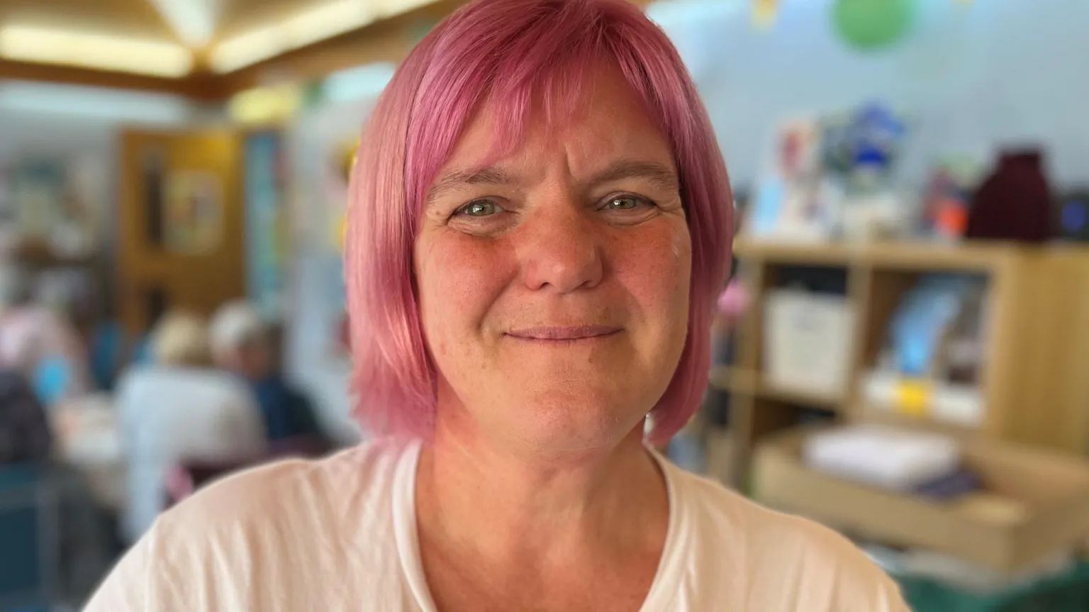 Close-up portrait of Karen McCurry with short pink hair wearing a white top, standing indoors with shelves and blurred figures in the background.