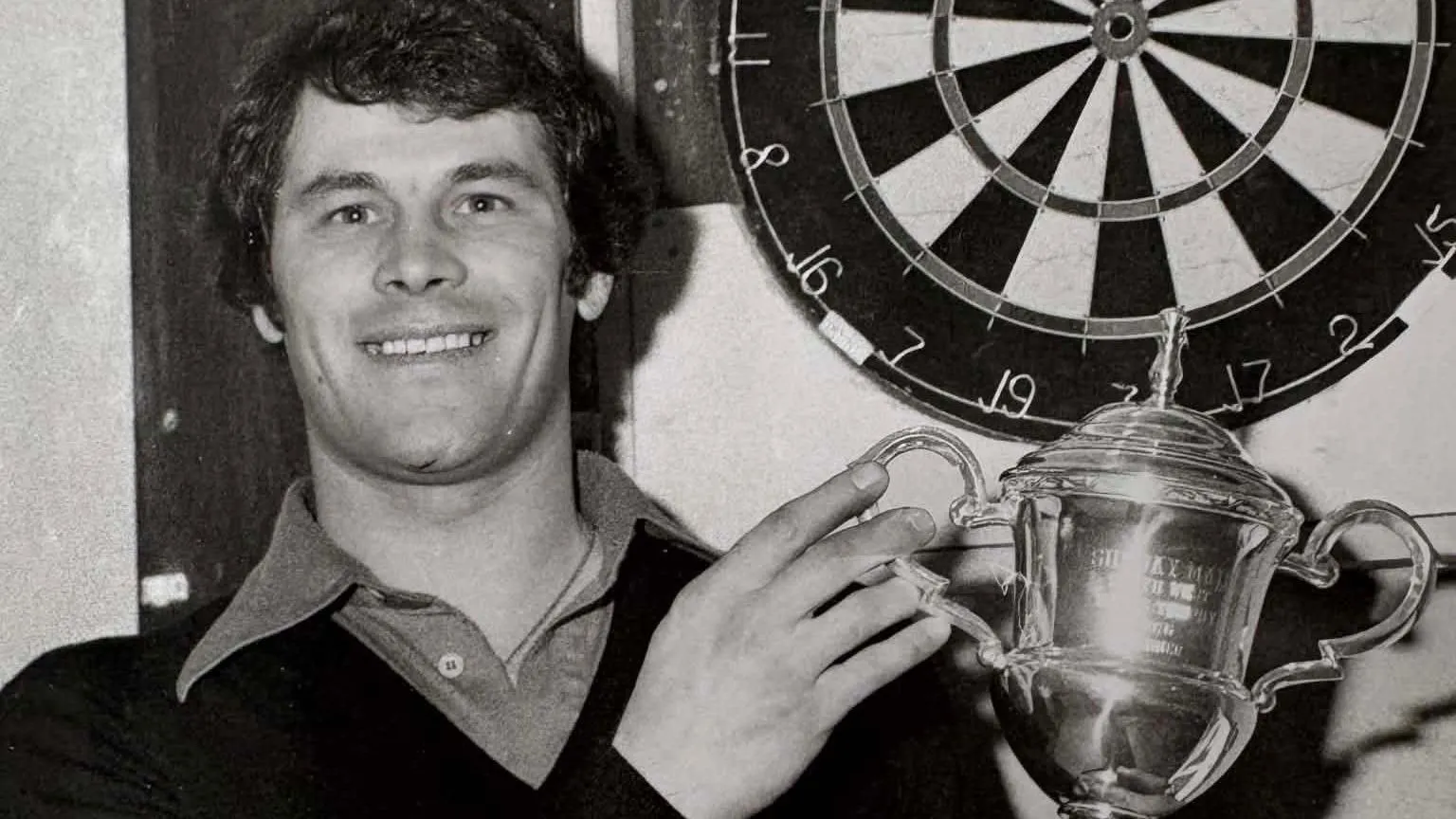 Smith Family A black and white picture of Rab Smith, a man with dark hair in a dark jumper with a lighter shirt underneath. He is holding a trophy while standing in front of a dart board.