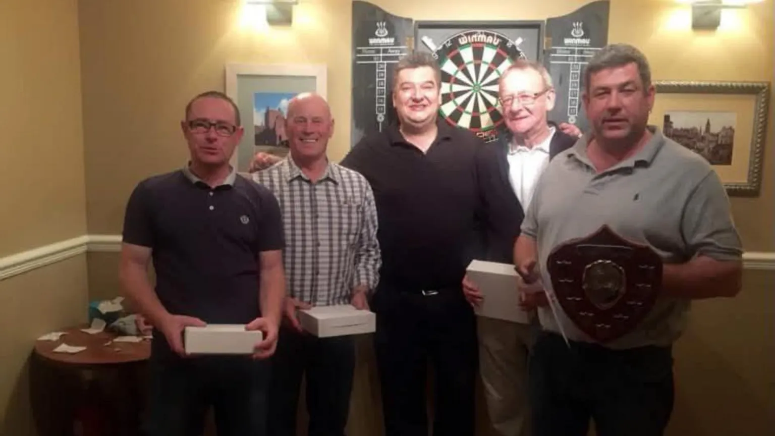Graham Lennox A group of men holding boxes and one with a trophy stand in front of a dart board