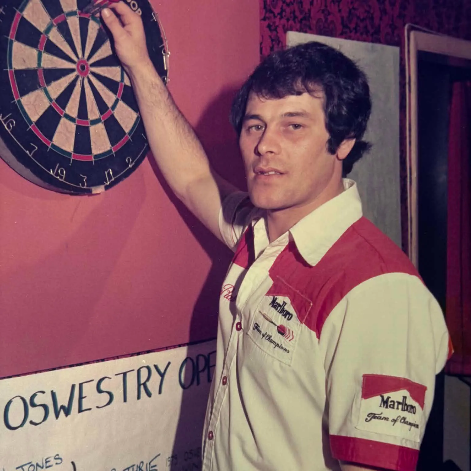  Rab Smith stands at a dart board with a white shirt with red shoulders. He has dark hair and is picking his darts out of the board
