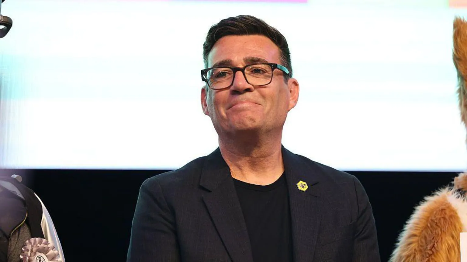  Burnham smiling, reacting to his victory in the Makerfield by-election. He is wearing a black top, black blazer and black framed glasses. He has dark brown hair.