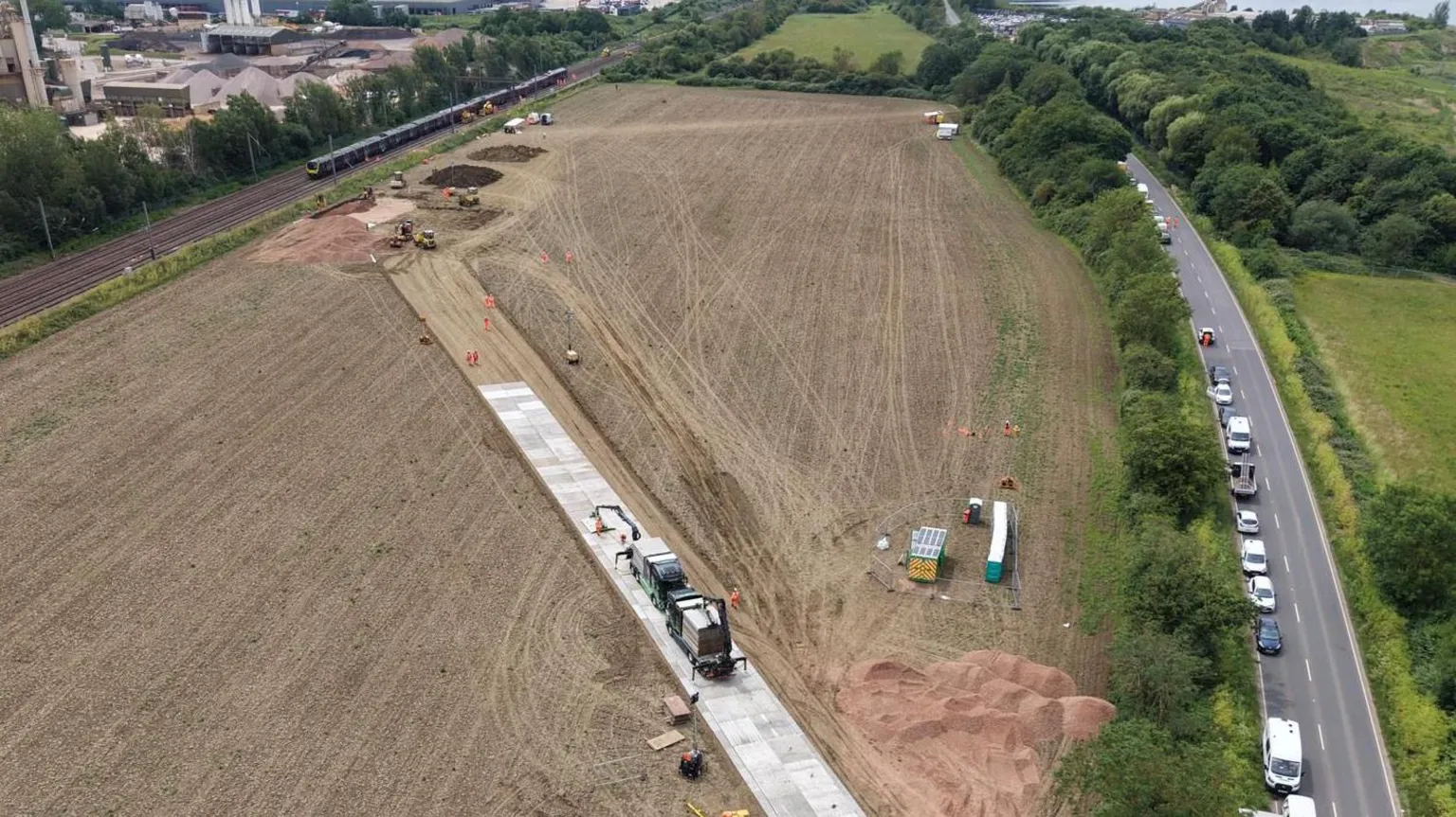 Network Rail Aerial view of a large brown field with a straight temporary concrete roadway running diagonally across it. It connects a road and the railway on either side of the field.
