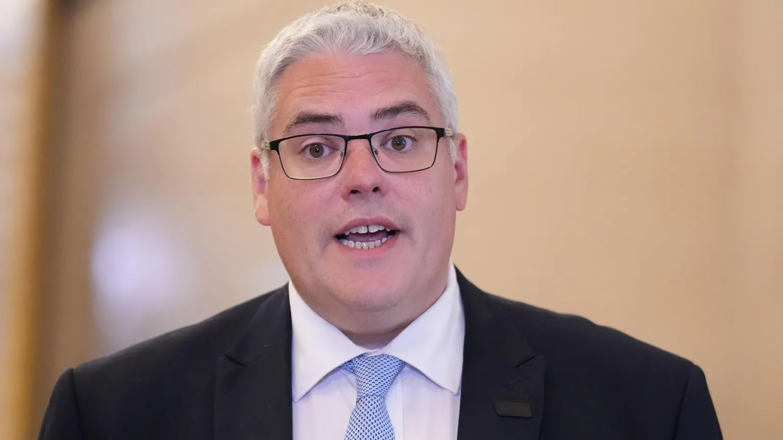 avin Robinson speaking. He has grey hair and is wearing black framed glasses, a black suit jacket, a white shirt and blue tie.